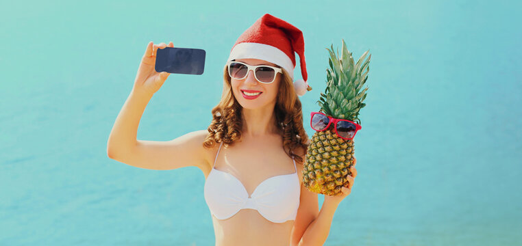 Christmas portrait of happy young smiling woman taking selfie picture by smartphone in red santa hat with pineapple over blue sea background