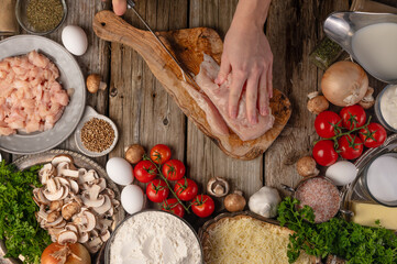 Chef hands cuts with knife chicken fillet for preparing meat filling on wooden table with variety of ingredients background. Concept of cooking process. Backstage of preparing pie. View from above.