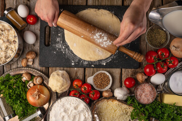 Chef hands rolls the dough with a rolling pin on wooden table with variety of ingredients background. Concept of cooking process. Backstage of cooking pie. View from above.