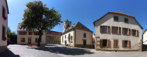 A medieval church and residential houses with half-hip roofs next to a square with a village tree in Rehborn center in Germany