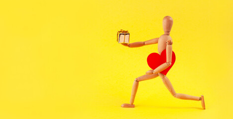 Yellow illuminating background and on it a wooden man with a red heart under his arm and a gift in his hand.
