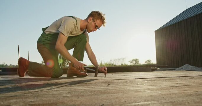 Young builder or workman dressed in dungarees kneeling down hammering in nails into wooden exterior flooring or deck in a low angle view