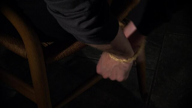Man struggles with hands bound behind back to wooden chair inside a dark room 