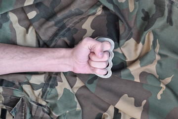 Male fist with brass knuckles on the background of a camouflage jacket. The concept of skinhead culture, handmade melee weapons