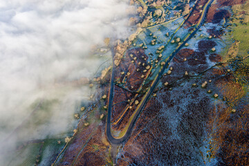 Aerial view of a narrow, winding mountain road emerging through a bank of fog and low cloud on a...