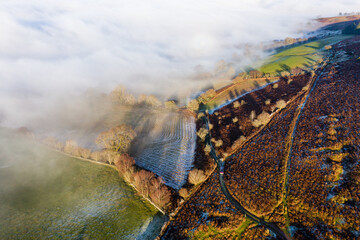 Aerial view of a narrow, winding mountain road emerging through a bank of fog and low cloud on a...