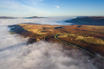 Aerial view of a narrow, winding mountain road emerging through a bank of fog and low cloud on a cold, frosty day