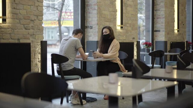 Millennial Caucasian couple surfing Internet on smartphones waiting for order in cafe on Covid-19 pandemic. Young man and woman in face masks messaging online in restaurant. Lifestyle concept.