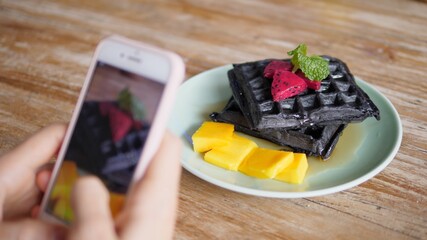 Female Food Blogger Taking Photo Of Black Vegan Waffles With Fruits Using Smartphone. 