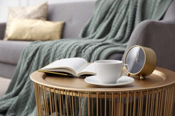Coffee table with cup of tea, clock and book near sofa in room