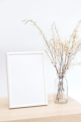Mock up of a white photo frame in a minimalistic interior with a bouquet of pampas grass in a glass vase against a white wall background