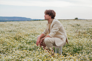 Naklejka na ściany i meble Tall handsome man sitting on a back of a chair in camomile flowers field
