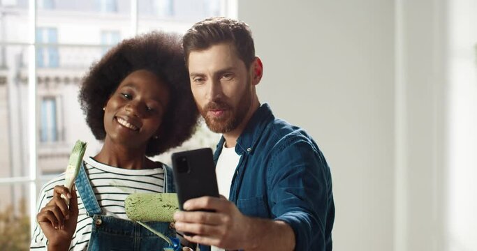 Portrait of happy mixed-races young married family couple man and woman smiling in new apartment taking selfie photos on smartphone posing with brushes in room. Home repair concept