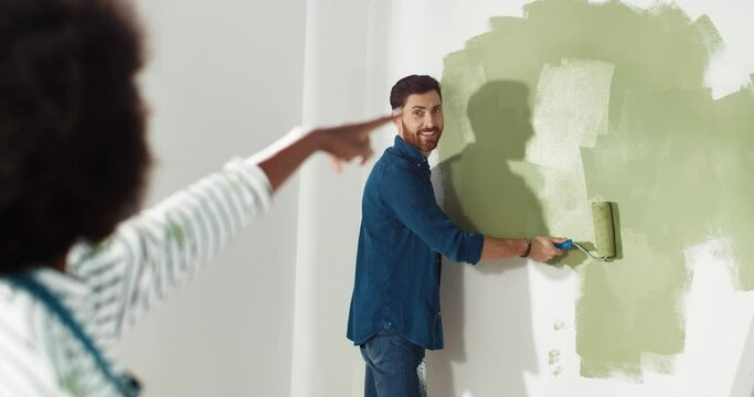 Rear of African American woman standing in home and showing her Caucasian husband how to repair and redecorate room. Man painting wall with brush roller and smiling. Renewing flat concept