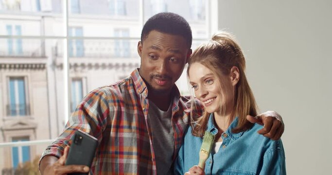 Close up portrait of happy multi-ethnic young family couple man and woman smiling in new apartment taking selfie photos on cellphone posing with brushes in room. Home repair. Remodeling concept
