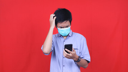 Asian man dizzy and stressed wearing mask looking at the cellphone isolated red background