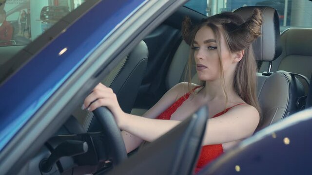 Portrait of beautiful young girl in red underwear sitting behind wheel in expensive car in dealership. Female with luxurious sport vehicle in motor show and auto business