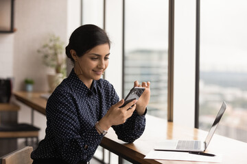 Happy young Indian woman look at smartphone screen texting or messaging online on gadget. Smiling millennial female user or client use modern cellphone at workplace in office. Technology concept.