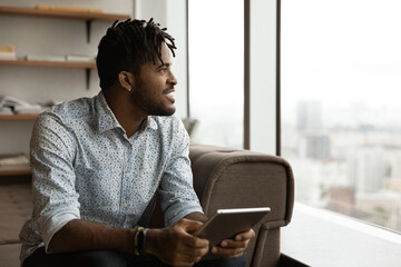 Smiling biracial man relax at home use modern tablet look in distance thinking or dreaming or...