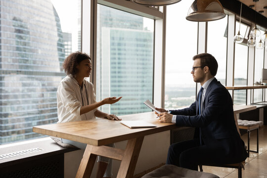 Multiracial businesspeople sit at desk in modern office brainstorm discuss business idea together. Male boss talk with biracial female candidate applicant at job interview. Employment, hiring concept. - Powered by Adobe