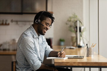Smiling millennial African American man in headphones watch online webinar on laptop making notes. Happy young biracial male in earphones take distant course or study on computer. Education concept.