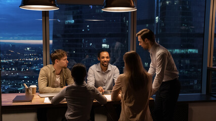 Wide banner panoramic view of multiracial colleagues work in office at night, discuss company strategies in group. Happy diverse multiethnic employees busy at workplace late hours. Teamwork concept.