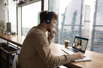 Smiling young Caucasian man in earphones talk on video call with colleague or client. Millennial...