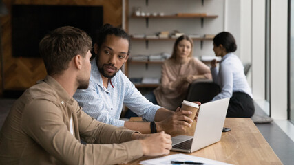Wide banner view of multiracial employee sit at desk in office work on laptop together discuss project. Focused young diverse businesspeople cooperate on computer brainstorm at meeting in office.