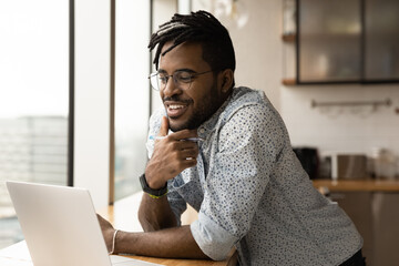 Smiling young African American man look at computer screen watch webinar on gadget make notes. Happy millennial biracial male work online or study distant on laptop in home office. Technology concept.