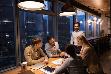 Diverse employees have late office group meeting discuss company financial paperwork together. Multiracial colleagues coworkers work analyze business ideas in boardroom at night, meet deadline.