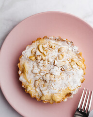 Delicious apple tartlet with caramelized apple, custard cream, roasted nuts and powdered sugar served on a pink plate with light background. Selective focus. Copy space.