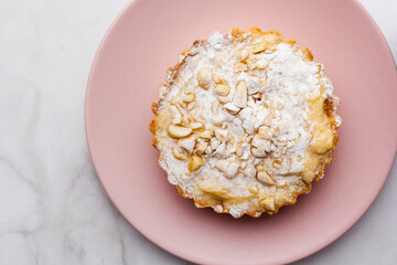 Delicious apple tartlet with caramelized apple, custard cream, roasted nuts and powdered sugar served on a pink plate with light background. Selective focus. Copy space.