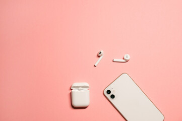 Simple flat lay with a stylish white smartphone and modern wireless earphones with a charging case on a pastel pink background.