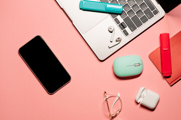 Flat lay on a pastel pink background with a silver laptop computer, smartphone with a touch screen, wireless mouse, earphones, stylish goggles, colorful markers, and notebook.