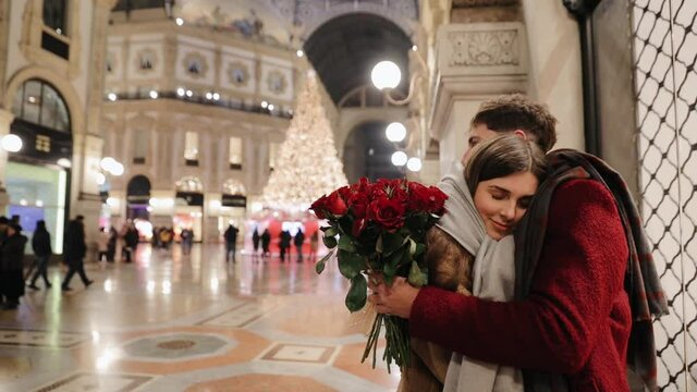 The young man hugging his girlfriend after engagement proposal in the mall on background of Christmas tree. Romantic smiling couple in love celebrating some date of their relationship with flowers