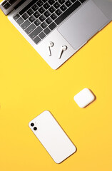 Vertical flat lay composition on a yellow background with a silver grey laptop, white mobile phone, and wireless earphones with a charging case.