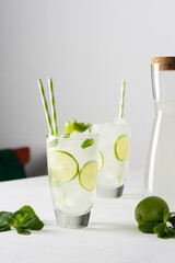 Refreshing cold drinks with lime and mint, glasses with lemonade and ice and straws, jug with drink on white background