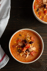 Spicy mexican soup with beans and corn, tomatoes and chicken breast, bowl of soup and napkin on dark wooden background, top view
