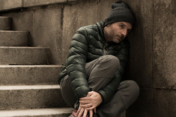 dark and edgy urban portrait of middle aged sad and depressed unemployed man sitting outdoors on...