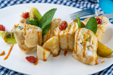 Simple homemade pancakes cut with banana, kiwi, strawberry jam on a white plate. Creamy curd filling. The food is decorated with mint. Rolled thin roll of pancake.