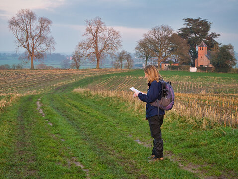 A single woman walker in walking gear on a route through a cut field of crop stubble checks her route with a map. Taken in Cheshire, UK at dusk on a calm overcast day in winter.