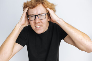 Red-haired man with glasses holding his head on a white background in a black T-shirt