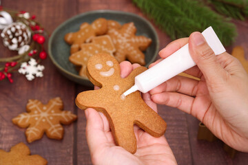 Obraz na płótnie Canvas A baked of cut out Christmas sugar biscuit in a gingerbread man shape in hands while decorating with royal icing from a piping tube. With various shapes of cooked cookies in background 