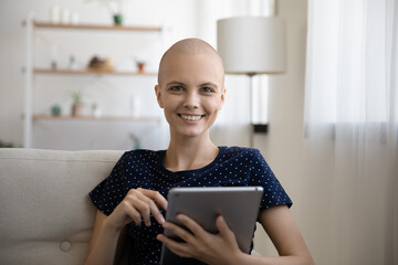 Life continues. Portrait of active woman recovering from oncology posing on couch at home holding pad device. Happy young female sick with cancer look at camera chat online with friend using tablet pc