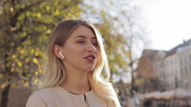 Attractive young blonde female with red lips in the city speaking in headphone. close up portrait happy beautiful female walking in European city. Smiling woman uses a earphones, headset.
