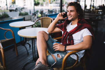 Cheerful handsome male laughing during smartphone conversation on free time holding coffee cup, positive young millennial man in casual wear smiling making mobile phone in roaming on cafe terrace