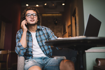 Handsome male millennial in trendy eyewear for vision correction talking on mobile phone during remote job, young curly hipster guy working distantly using laptop computer and making cellular call