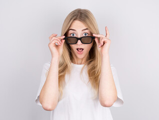Portrait of a surprised young beautiful girl in eyeglasses looking at camera while raised up glasses. Isolated over gray background