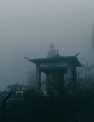 A tall statue of Lord Buddha near a buddhist temple, standing in middle of dense winter fog in morning at Lolegaon, Kalimpong. 