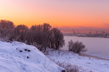 A soothing winter landscape at sunset with frost-covered trees on the banks of a frozen river.